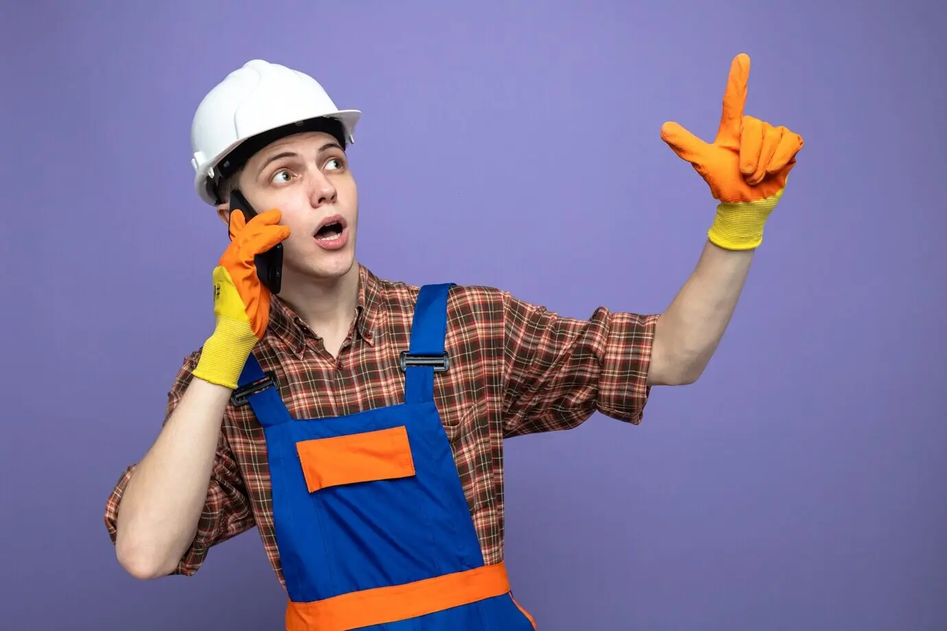 Überraschter junger männlicher Bauarbeiter in Uniform mit Handschuhen zeigt nach oben und spricht am Telefon.